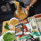 A person holds a large metal bowl filled with stir-fried noodles above a spread of ingredients and packaged food on a dark kitchen counter. Visible items include a package labeled 'Soyumaki', a bowl of chopped green onions, parsley, garlic, and various seasoning containers.